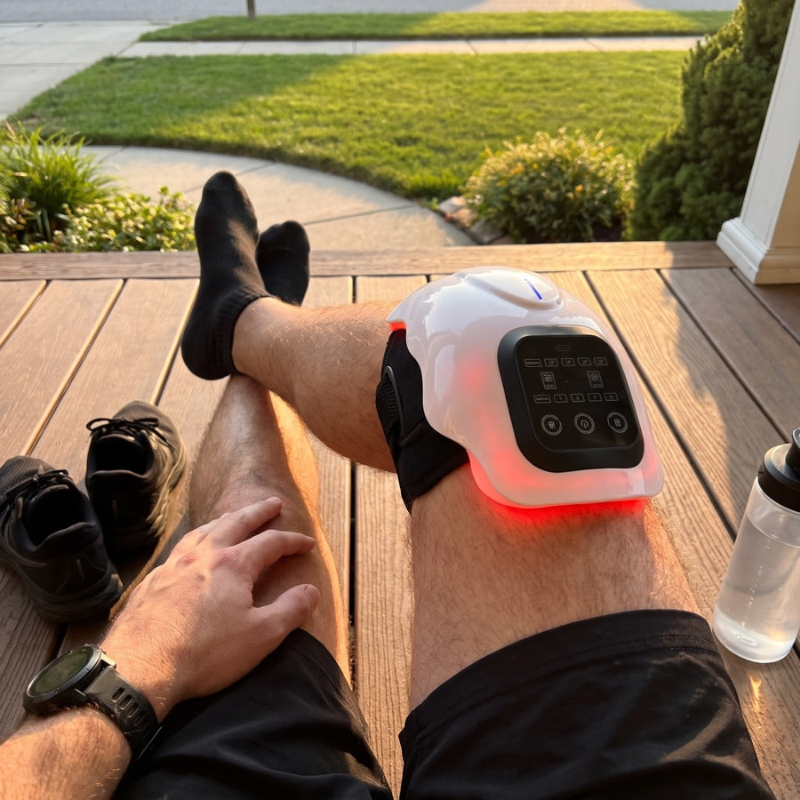 A man resting on a deck with the massager wrapped around his knee, its red light showing